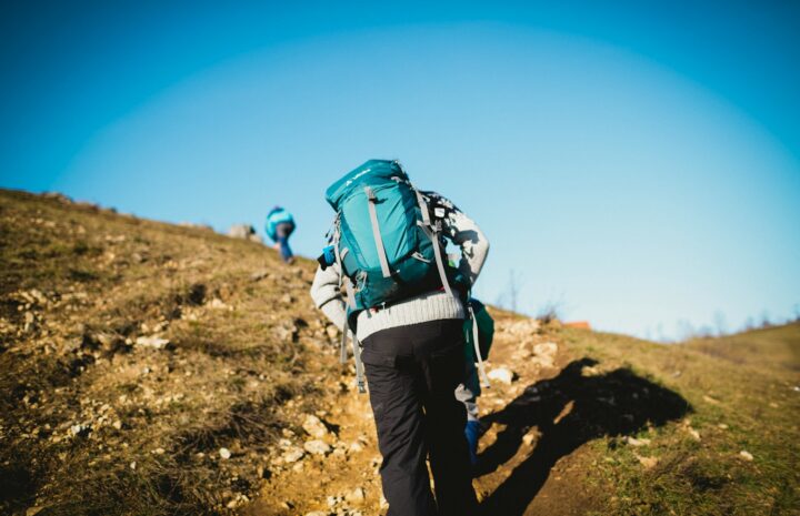 man in blue jacket and black pants with blue backpack walking on brown field during daytime